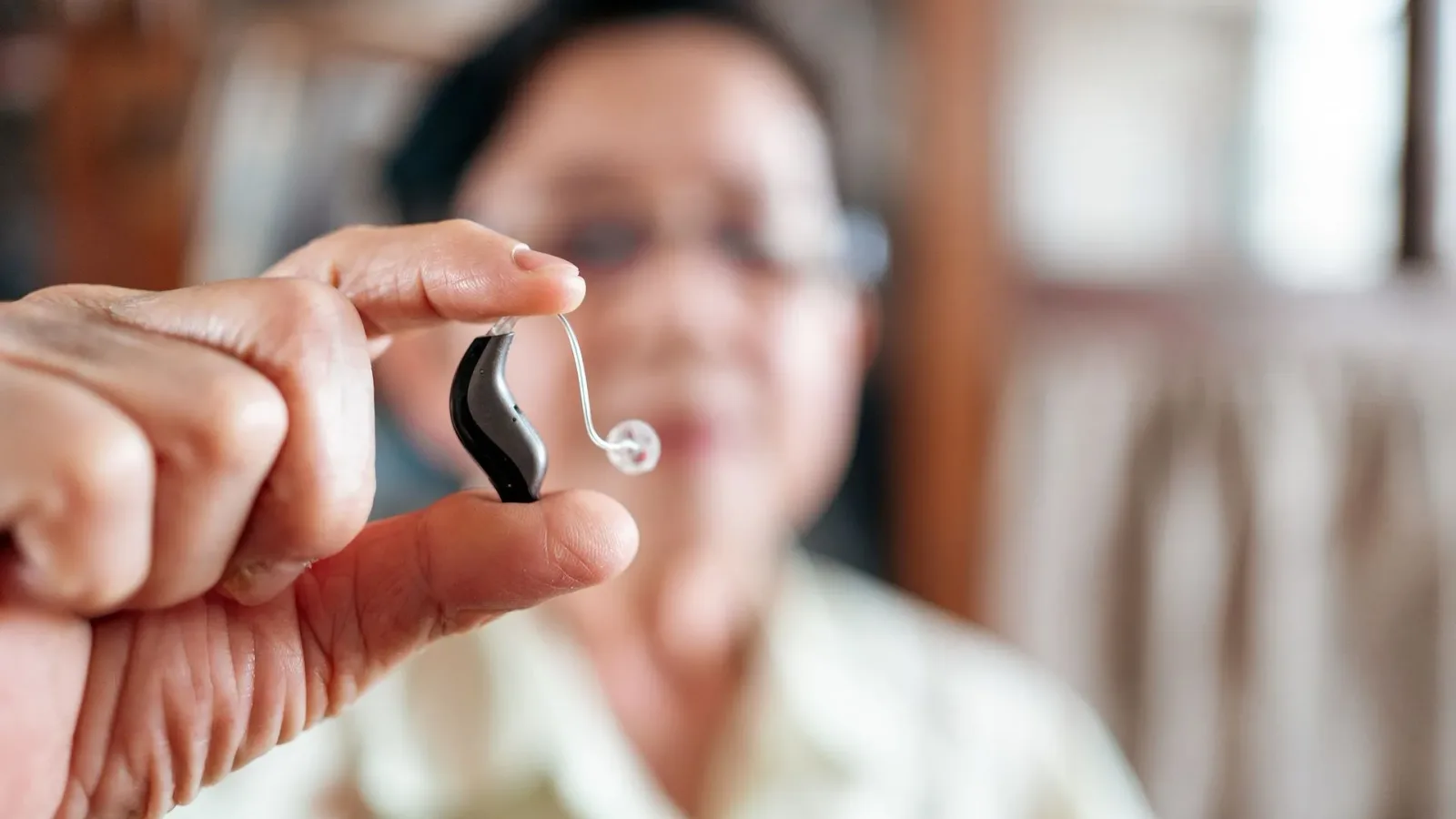 Close-up of a hand holding a small behind-the-ear hearing aid, with a smiling older adult in soft focus behind it