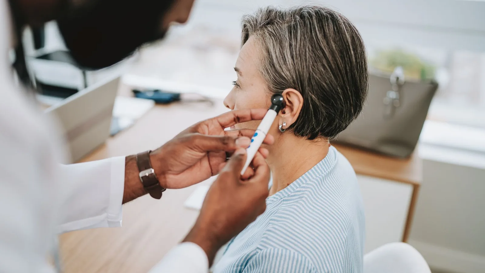 An audiologist gently examines a patient's ear with an otoscope in a bright, modern clinic